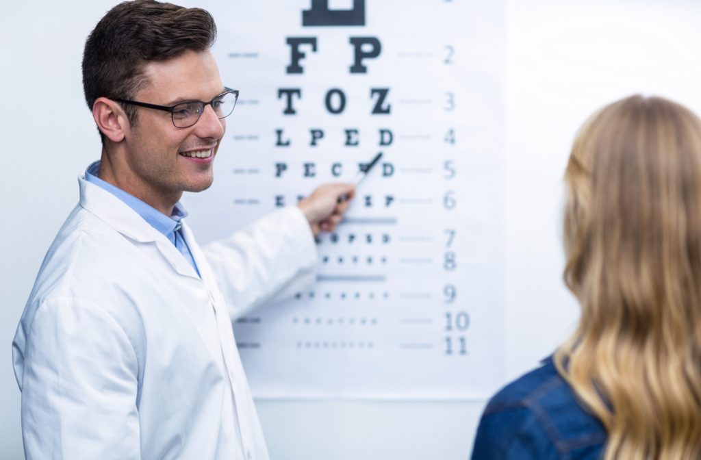A smiling optometrist points to an eye chart while a female patient faces towards it.