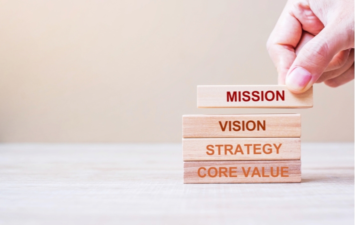 Image of a hand stacking wooden blocks with the words Mission, Vision, and Strategy, with Core Value supporting the stack.